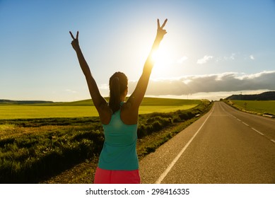 Woman Celebrating Running And Training Success On Countryside Road During Sunset Or Sunrise. Female Runner Raising Arms Towards The Sun.