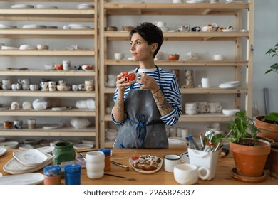 Woman ceramist drinks tea with bun in ceramics studio. Coffee break after pottery master class or work in handicraft shop. Female artist stands against shelving with handmade utensil - Powered by Shutterstock