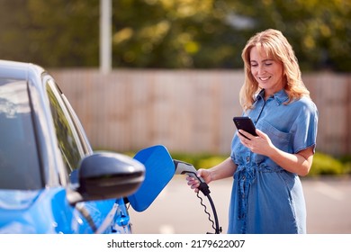 Woman Charging Electric Car With Cable Using App On Phone To Monitor Battery Level