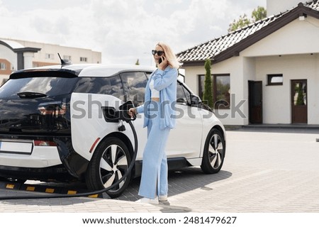 Woman Charging Electric Car While Talking On Phone In Front Of Modern House