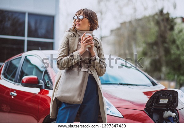 Woman charging electro car at the electric gas
station and drinking
coffee