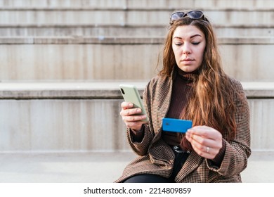 Woman In Checkered Coat Entering Credit Card Credentials Into Cellphone While Sitting On Stairs And Making Online Order On City Street