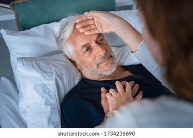 Woman Checking Fever Temperature Of Senior Man Lying On Bed. Mature Husband Resting At Home Feeling Symptoms Of The Flu While Wife Checking Fever By Touching Forehead. Close Up Face Of Old Sick Man.