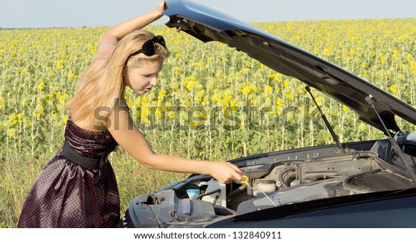 Woman checking the\
level of the engine oil in her car which has broken down alongside\
a field of sunflowers