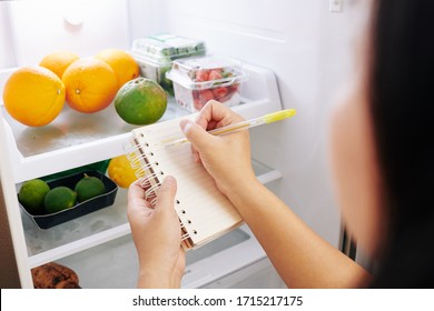 Woman checking refrigerator and making shopping list before going to grocery store