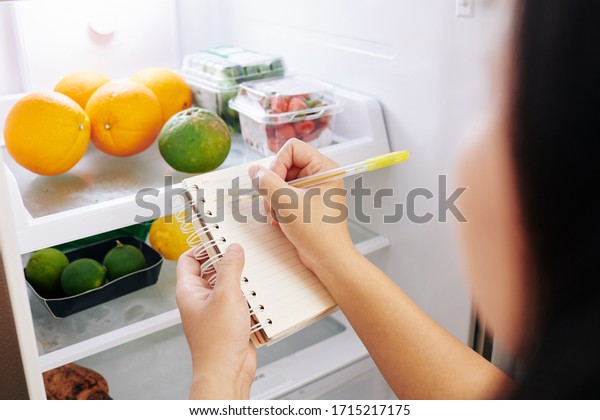 Woman checking refrigerator and making\
shopping list before going to grocery\
store