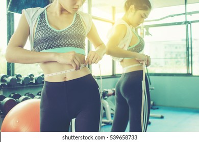 Woman Checking Success Of Weight Loss Program With Measuring Tape In Front Of Mirror At Gym.