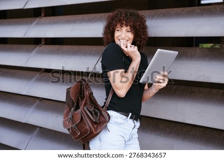 Woman checking tablet while standing by louver wall, backpack on shoulder, focused on emails and schedule, remote work vibe, compact urban backdrop, confident