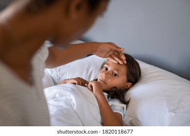 Woman Checking Temperature With Hand Of Little Ill Daughter. Mother Checking Temperature Of Her Sick Indian Girl. Sick Child Lying On Bed Under Blanket With Woman Checking Fever On Forehead By Hand.