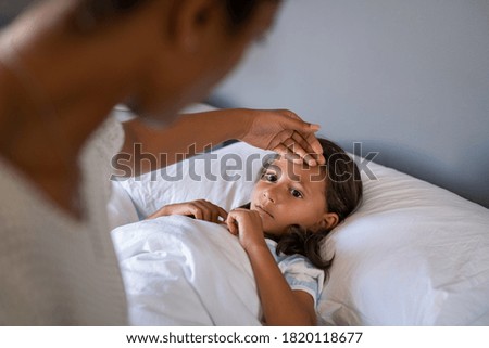Woman checking temperature with hand of little ill daughter. Mother checking temperature of her sick indian girl. Sick child lying on bed under blanket with woman checking fever on forehead by hand.