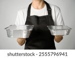 Woman chef holding a aluminum lunch box with food, close-up. Female chef in apron holding foil lunch box, white background. Takeaway food delivery in restaurant.