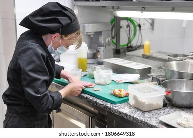 Woman Chef In Protective Face Mask Prepare Food In The Kitchen Of A Restaurant Or Hotel. Coronavirus Prevention Concept. High Quality Photo