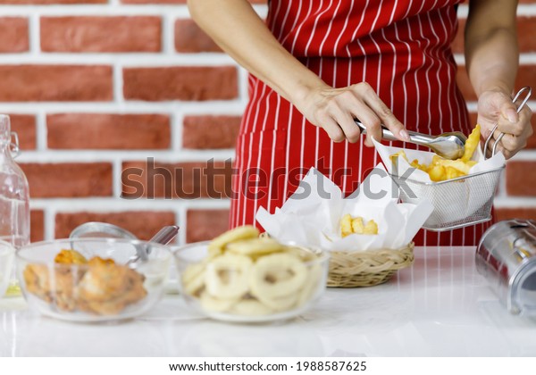 Woman chef in red striped apron grip tasty fried\
potatoes to share them from metal sieve to wicker basket on cooking\
table near brick wall in home kitchen to arrange food on dishes\
before serving