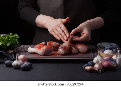 Woman chef rubs her paprika with fresh raw chicken drumsticks on a dark background. Nearby lie the ingredients for cooking: shallots, blue plums, garlic pepper, salt and parsley. - Shutterstock ID 708297244