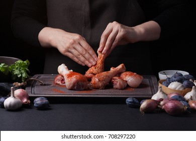 Woman chef rubs salt with fresh raw chicken drumsticks on a dark background. Nearby lie the ingredients for cooking: shallots, blue plums, garlic pepper, salt and parsley. - Shutterstock ID 708297220