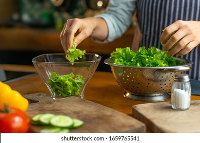 A woman chef in a striped apron is is tearing freshly washed, fresh, clean leaves of lettuce into a separate glass bowl. She is located in a rustic style kitchen. - Powered by Shutterstock