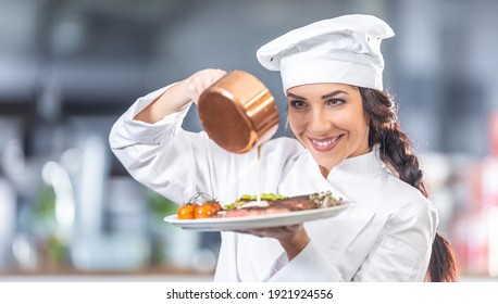 Woman in chef's uniform pours sauce over steak with vegetables in the kitchen.