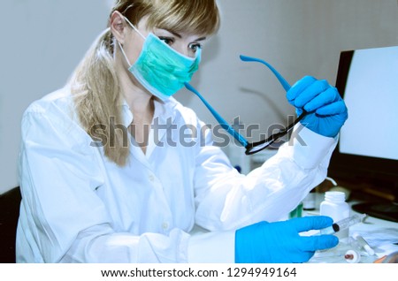 Woman chemist in clinic office working on new cure holding syringe in hand.Looking thoughtful, preparing to research new sample wearing steril mask and latex gloves.Put eye glasses to proceed a work 