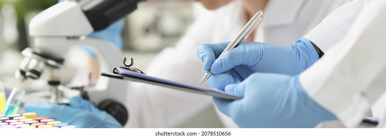 Woman chemist looking through microscope in laboratory. Assistant writing data to paper. Laboratory diagnostics of diseases concept - Shutterstock ID 2078510656