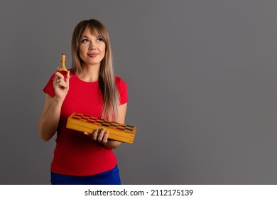 Woman Chess Player Holding Chess Piece White King And Chessboard. Girl In Red T-shirt Dreamily Looks Up While Standing On Gray Background. Concept Of Study Chess, Women In Chess Sport. Self Education