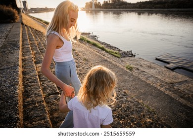 A woman and a child enjoy a serene summer evening walk by the banks of the Drava River in Osijek, Croatia, as the sun sets and casts a warm glow over the water. - Powered by Shutterstock