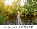 A woman and child walk across a narrow suspension bridge over a calm river, surrounded by lush green forest, capturing a serene moment in nature