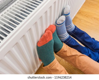 Woman and child wearing colorful pair of woolly socks warming cold feet in front of heating radiator in winter time. Electric or gas heater at home. Part of body, selective focus.  - Shutterstock ID 1196238214