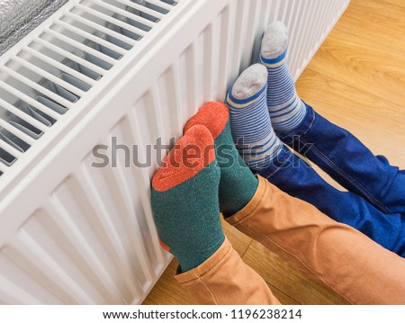 Woman and child wearing colorful pair of woolly socks warming cold feet in front of heating radiator in winter time. Electric or gas heater at home. Part of body, selective focus. 