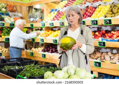 Woman Choosing Melon While Standing In Greengrocer. Old Man Shopping In Background.