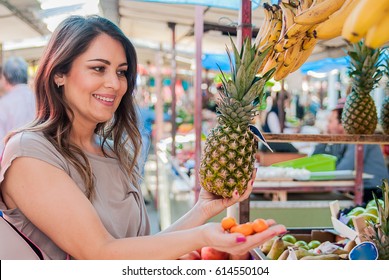 Woman Choosing Pineapple During Shopping At Fruit Vegetable Green Market. Attractive Woman Shopping. Beautiful Young Woman Picking Up, Choosing Fruits, Pineapples.