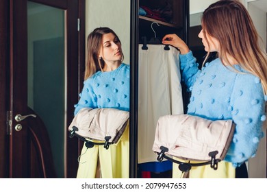 Woman Choosing Trousers And Skirts On Hangers For Outfit Wearing Warm Clothes By Wardrobe Mirror. Girl Holding Pants In Closet At Home. Fashion Choice