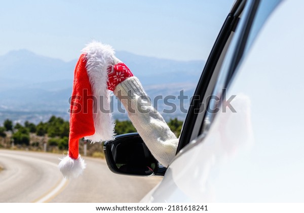 Woman in christmas gloves driving a car\
holding santa claus hat from the open window. Trip on the\
serpentine road in the\
mountains.