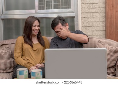 A woman chuckles at her cowardly husband covering his eyes while watching a scary movie on their laptop. Casual and lighthearted scene at the living room. - Shutterstock ID 2256865443