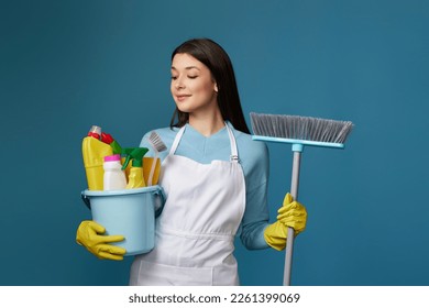 woman in cleaner apron holding bucket of detergents and broom