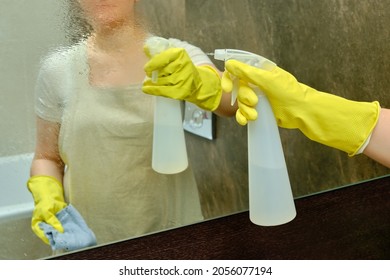 A woman is cleaning the bathroom by wiping the mirror with a rag. Disinfection with yellow rubber gloves in the washroom - Powered by Shutterstock