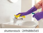 Woman cleaning the bathroom sink, she is polishing the faucet using a sponge wipe and spray detergent