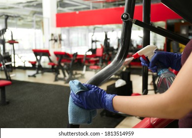 Woman Cleaning Exercise Equipment With Disinfectant Spray And Cloth In Gym, Closeup