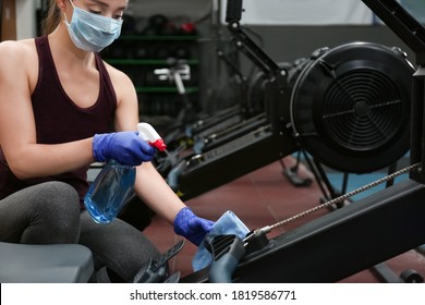 Woman Cleaning Exercise Equipment With Disinfectant Spray And Cloth In Gym