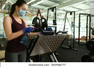 Woman Cleaning Exercise Equipment With Disinfectant Spray And Cloth In Gym