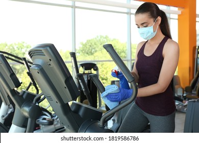 Woman Cleaning Exercise Equipment With Disinfectant Spray And Cloth In Gym