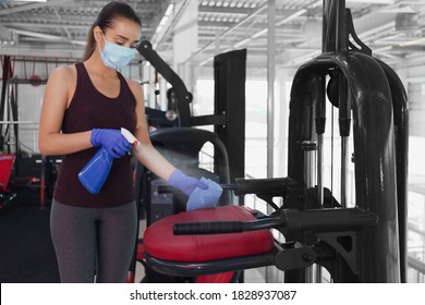 Woman Cleaning Exercise Equipment With Disinfectant Spray And Cloth In Gym