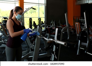 Woman Cleaning Exercise Equipment With Disinfectant Spray And Cloth In Gym