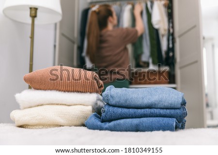 A woman is cleaning out her wardrobe, standing by clothes placed on hangers and in drawers. In the foreground are stack of sweaters and jeans