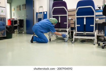 Woman From The Cleaning Service Of A Hospital Crouching Cleaning The Wheels Of A Patient Chair In The Operating Room Beforehand. Sanitary Concept