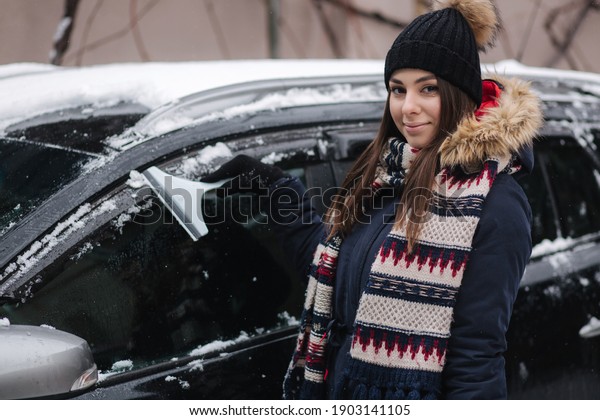 A woman is cleaning snowy window on a\
car with snow scraper. Pretty woman warmy dressed clean her car\
outdoors. Cold snowy and frosty morning. Black\
car