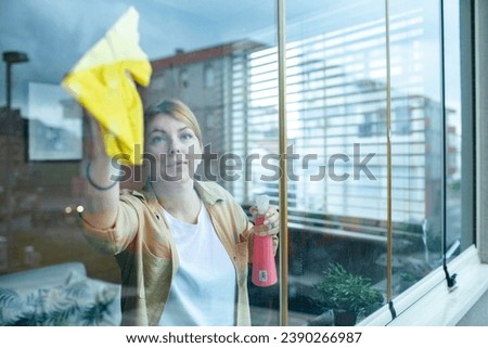 Woman cleaning window with cleanser sprayer and yellow rag at home