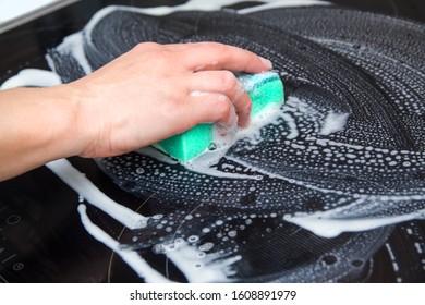 A woman cleans the electronic ceramic hob by green sponge with special detergent agent applied to it. Chores and domestic labour concept.