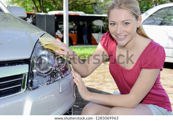 Woman cleans headlight of car with cleaner and\
chamois on a sunny day