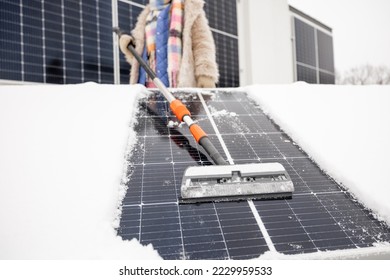 Woman cleans solar panels from snow to produce power in winter on the roof of her house. Energy independence and sustainability concept. View from a bottom focused on swab - Shutterstock ID 2229959533