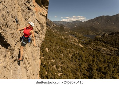 A woman is climbing a rock face with a red tank top on. The mountain range in the background is lush and green - Powered by Shutterstock